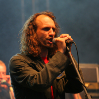 At the 2011 Kilkim Žaibu festival, a man with long curly hair performs on stage, passionately singing into a microphone he holds with both hands. He wears a dark jacket over a red shirt and has a lanyard around his neck. The background is dark with heavy stage lighting and another person, partially visible, is in the background.