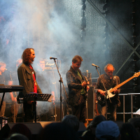 A band performs on an outdoor stage at the 2011 Kilkim Žaibu festival, illuminated by soft lighting and surrounded by mist. Five musicians play keyboard, guitar, bass, and percussion. All are dressed in casual outfits and jackets. A backdrop displays a faint image while a crowd watches.