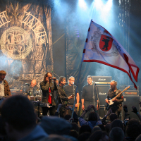 A rock band performs on a well-lit stage with a large crowd in front. Seven band members stand with instruments, including guitars and a drum set. A large backdrop with intricate artwork and text is behind them, reminiscent of Kilkim Žaibu 2011. The crowd waves a flag bearing a crest with a red center and a black figure.