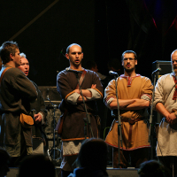 A group of five men dressed in medieval-style clothing stands in a line on a dark stage at the 2011 Kilkim Žaibu music festival, each positioned in front of a microphone. They appear focused, possibly singing or speaking to the audience. Musical equipment is visible in the background, indicating a live performance setting.