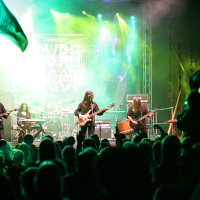 A rock band performs energetically on a vibrant stage, illuminated by green and purple lights at the 2011 Kilkim Žaibu festival. The setup includes guitars, a drum set, and keyboards. Enthusiastic fans with raised flags and hands gather closely in front of the stage, enjoying the live music experience.