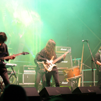 A rock band performs on stage at the 2011 Kilkim Žaibu festival with three guitarists positioned under green stage lights. Each musician is playing an electric guitar or bass while dressed in dark clothing. Amplifiers and other band equipment are visible in the background, contributing to the energetic atmosphere.