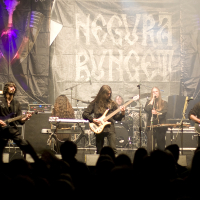 Band performing live on stage with five members playing instruments: two guitarists, one keyboardist, a drummer, and a bass guitarist in the center. The backdrop displays the band's name, "Negură Bunget". The scene includes amplifiers and stage lighting at Kilkim Žaibu 2011, while a crowd watches in the foreground.