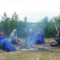 A group of people, some wearing hooded jackets and blankets, sit around a campfire in a grassy field during Kilkim Žaibu 2011. They appear to be engaging in conversation and staying warm. Logs encircle the fire, and trees are visible in the background on a cloudy day. Smoke from the fire rises into the air.