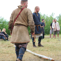 Two men dressed in historical medieval attire participate in an outdoor event, reminiscent of Viking-era clothing. Set in a grassy, open area during Kilkim Žaibu 2011, one man walks past a fallen wooden branch while others linger in the background. Spectators are seated comfortably in folding chairs.