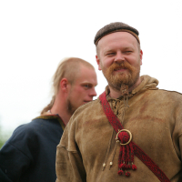 Two men in Viking-era clothing stand outdoors at Kilkim Žaibu 2011. The man in the foreground wears a brown tunic, a small hat, and a red sash with a circular brooch. He has a beard and smiles at the camera, while the man in the background, with a short beard and dark tunic, looks away to the left.