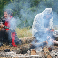 Two people sit on log benches around a smoky campfire at the 2011 Kilkim Žaibu festival. The person on the left wears a black coat and red scarf, holding a drink, smiling. The person on the right is dressed in a white rain poncho and patterned pants, also holding a drink and looking down. The background is grassy and wooded.