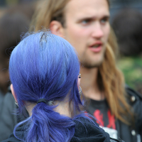 A person with long, vibrant blue hair tied in a ponytail is in the foreground, facing away. In the background, slightly out of focus, stands another with long blonde hair and a beard. Both are outdoors at Kilkim Žaibu 2011, wearing dark clothing. The scene suggests a casual or social setting.