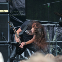 A long-haired musician intensely plays an electric guitar on stage at the 2011 Kilkim Žaibu festival. He is headbanging, with his hair swinging wildly. Behind him are Marshall amplifiers and a drum set. The audience is visible in the foreground, creating an energetic scene likely at a rock or metal concert.