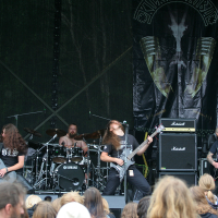 A metal band is performing on the outdoor stage of Kilkim Žaibu 2011, adorned with large, dark banners. Four members, dressed in black, are playing electric guitars and bass, with long hair flying as they headbang. The drummer is visible at the back, surrounded by Yamaha drums. The audience watches intently with some raised hands.