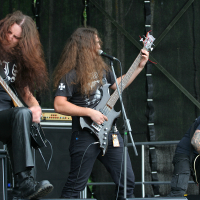 Three long-haired metal musicians perform energetically on stage at the 2011 Kilkim Žaibu festival. The lead singer, with both hands on his guitar, sings into a microphone, while the flanking guitarists headbang intensely. Amplifiers, including Marshall stacks, are visible in the background. The overall scene is dynamic and intense.