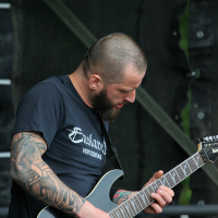 A bearded man with a shaved head and intricate tattoos on his left arm plays an electric guitar. Wearing a black t-shirt with white text, he is focused on his instrument. The background shows a dark, partially blurred stage setup, reminiscent of Kilkim Žaibu 2011.