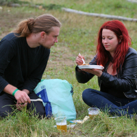 Two people, a man with long hair tied back and a woman with red hair, sit on grass sharing a meal at Kilkim Žaibu 2011. The woman, wearing a black leather jacket, holds a plate and fork while the man looks on. They have drinks in plastic cups on the ground next to them, along with a plastic bag and utensils.