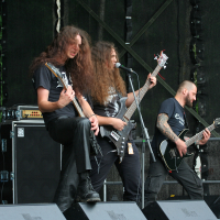 A heavy metal band performs on stage at the 2011 Kilkim Žaibu festival, with four male musicians playing electric guitars and bass. The vocalist in the center, with long hair, sings passionately into a microphone. Professional audio equipment, including Marshall amplifiers, is visible. All wear black clothing.