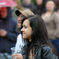A young woman with long dark hair and a black leather jacket stands in a crowd at Kilkim Žaibu, holding a plastic cup with a drink. She is smiling and looking to her left. The background is slightly blurred, showing several people and a red umbrella, indicating overcast weather.