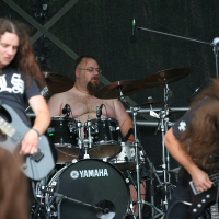 A rock band performs on stage at Kilkim Žaibu 2011. The drummer, shirtless, passionately plays on a Yamaha drum kit. Two guitarists with long hair are in the foreground, intensely engaged with their instruments, headbanging. The stage backdrop is dark, emphasizing the musicians' energetic presence.