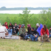 A group of people are seated in a semicircle around a campfire by a lakeside, reminiscent of the 2011 Kilkim Žaibu festival. They are dressed in casual, warm clothing, with many wearing hoodies. The background features lush green reeds and trees, with the lake visible behind. The sky is overcast, suggesting a cool day.