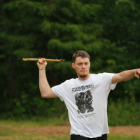 A person with short hair wearing a white graphic T-shirt and dark pants stands on grassy terrain, raising one arm to throw a stick and extending the other arm forward. There are trees in the blurred background, and the weather appears to be slightly misty or rainy, reminiscent of a scene from Kilkim Žaibu 2011 festival.