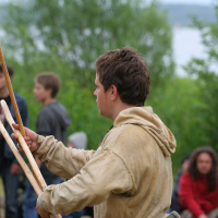 A person dressed in historical attire, including a beige tunic and hood, stands outdoors at what appears to be the 2011 Kilkim Žaibu festival while holding wooden sticks. They are in a green, forested area, with blurred figures and trees in the background. The weather appears cloudy or rainy. People in modern attire are observed.
