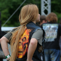 A person with long blonde hair is facing away from the camera at an outdoor event, possibly Kilkim Žaibu 2011. They are wearing a black denim vest featuring a graphic of a flaming skull and a grey t-shirt. A studded belt can be seen at their waist. Other event attendees are visible in the background.