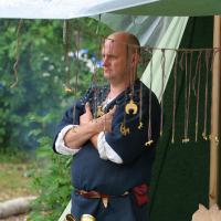 A person in medieval attire stands under a canopy with arms crossed. Various brass artifacts, including keys and coins, hang on strings in front. The table displays more artifacts on a black cloth. The setting appears to be the 2011 Kilkim Žaibu festival, with green foliage in the background.