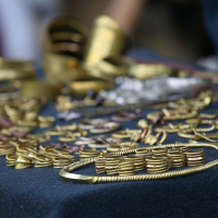 A close-up view of assorted gold jewelry pieces displayed on a dark cloth. The collection, reminiscent of the opulence seen at Kilkim Žaibu 2011, includes bracelets, rings, necklaces, and other ornamental items with intricate designs and metallic luster. Some pieces are arranged in rows; others are scattered, showcasing various sizes and shapes.