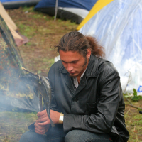 A man with a ponytail and wearing a black leather jacket crouches near a campfire at the 2011 Kilkim Žaibu festival, roasting two fish on a skewer. He is focused on the cooking. In the background, colorful camping tents are set up on grassy ground. Smoke from the campfire partially obscures his face.