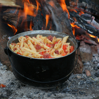 A black pot filled with pasta, sausage, and diced tomatoes sits over an open campfire at the 2011 Kilkim Žaibu festival. The flames and logs beneath the pot are visible, indicating the cooking process. The surrounding ground has ash and pieces of wood, evoking a rustic outdoor cooking experience.