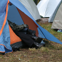 A blue and orange tent is set up on grassy ground. The tent flap is open, revealing three pairs of legs in black pants and black boots, partially covered by blankets. Other tents are set up in the background, suggesting a camping area at the 2011 Kilkim Žaibu festival with multiple tents.