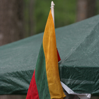 A tricolor flag with vertical stripes of yellow, green, and red is attached to a corner of a green tarp-covered structure. The flag's fabric is slightly creased, fluttering in the breeze at the Kilkim Žaibu festival in 2011. The background is blurred greenery with hints of tree trunks, suggesting an outdoor setting.