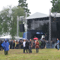 An outdoor music festival, Kilkim Žaibu 2011, features a stage with a canopy and equipment for amplification. A crowd gathers under overcast skies, some with umbrellas and rain ponchos. Trees frame the scene, creating a natural backdrop. A blue poncho stands out in the foreground amidst spectators.