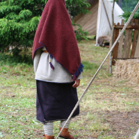 A person stands outdoors, covered head-to-toe in traditional clothing. They wear a deep red shawl over their head and shoulders, a white outer garment, a long dark skirt, and brown shoes. This scene from the 2011 Kilkim Žaibu festival features green trees, a rustic wooden fence, and a tent on grassy, slightly muddy ground.