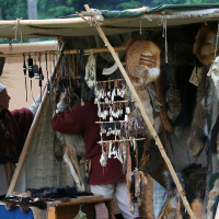 A market stall inside a canvas tent at 2011's Kilkim Žaibu festival displays various animal pelts and bone crafts. The setup features fur hides, animal skulls, and bone jewelry hanging on wooden beams. Two individuals, partly obscured, are arranging the items. The background shows a dense forest.