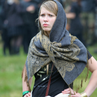 A young woman with blond hair stands outdoors in a grassy area during Kilkim Žaibu 2011. She wears a dark, intricately patterned shawl over her head and shoulders, a black shirt, and a white garment around her waist. With a serious expression, people can be seen in the blurred background under a green canopy.