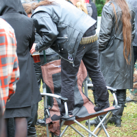 A person with long hair and a black leather jacket and pants stands on a folding camping chair at what appears to be the Kilkim Žaibu festival in 2011. The brown chair has armrests and a metal frame. Nearby, another person in a hooded jacket is visible, with more people in the background.