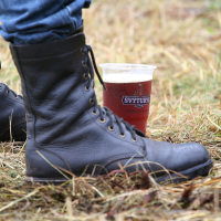 A close-up image of a person wearing black leather combat boots standing on grass at the Kilkim Žaibu festival. The individual is also clad in blue jeans. Beside the boots, there's a plastic cup filled with dark beer, crowned with white foam and bearing a visible label, capturing the essence of 2011 festivities.