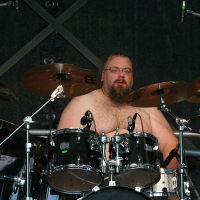A shirtless man with a beard plays a black drum set on stage, surrounded by cymbals. Holding drumsticks, he appears focused during the 2011 Kilkim Žaibu music festival. The background features multiple microphones and a dark backdrop, suggesting a live performance or concert setting.