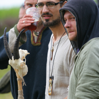 Three men are standing outdoors at Kilkim Žaibu 2011, dressed in medieval-inspired clothing. The man in the center has short dark hair, glasses, and a beard, and holds a staff adorned with a goat skull and horns. The man on the left is drinking from a cup, while the man on the right wears a hooded garment and has a light beard.