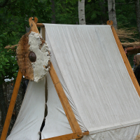 A small triangular canvas tent with wooden supports is set up in a forested area, reminiscent of the Kilkim Žaibu 2011 festival. A round shield with fur and leather adorns the left side of the tent. The background includes trees and partial glimpses of other tents or structures among the greenery.