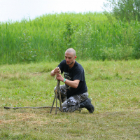A person wearing a black t-shirt and camouflage pants is kneeling on the grass, constructing a tripod structure with three wooden sticks. They are outdoors in a grassy, bushy area by a lake or river, reminiscent of the scenes from Kilkim Žaibu 2011.