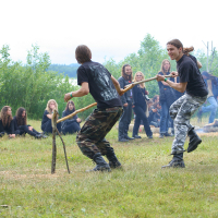 Two individuals engaged in a mock spear fight using wooden sticks, wearing black shirts and camouflage pants, while a group of spectators dressed in dark clothing sit and stand around a campfire in the background on a grassy field bordered by trees during the 2011 Kilkim Žaibu festival. The scene has a casual and relaxed outdoor atmosphere.