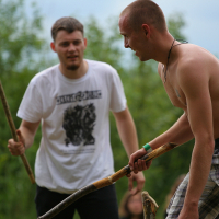 Two men engage in outdoor stick fighting at the 2011 Kilkim Žaibu festival. The shirtless man on the right holds a continuously swinging stick and faces off against a man in a white t-shirt. They stand in a grassy area with trees, where blurred foliage emphasizes the action in the foreground.