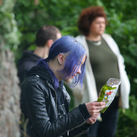 A person with blue hair and wearing a black leather jacket sits outdoors at Kilkim Žaibu 2011, intently reading the back of a snack package. Two other individuals, one wearing a white jacket and another in a black hoodie, are blurred in the background amidst green foliage.