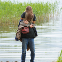 A person with long hair and wearing a black shirt and jeans stands on sandy terrain at the edge of a body of water, carrying another individual with a brown jacket and red handbag on their back, reminiscent of the spirited vibe at Kilkim Žaibu 2011. Tall green reeds and calm water are visible in the background.