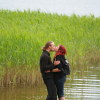 A couple stands in shallow water near the edge of a lake, surrounded by tall green reeds. They are kissing, with the man wearing a black long-sleeve shirt and pants, and the woman wearing a black top and dark shorts. Both have their arms around each other as if they were at Kilkim Žaibu festival. The man's brown boots rest on the sandy shore.