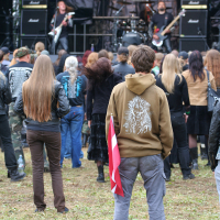 Outdoor concert at Kilkim Žaibu with attendees watching a live band perform on stage. One person in the foreground, wearing a brown hoodie, holds a red and white flag and stands out among the crowd. The stage, reminiscent of 2011 festivals, features musicians playing guitars and is flanked by large black speakers. The atmosphere is casual and festive.