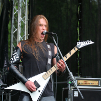 At the 2011 Kilkim Žaibu festival, a man with long hair passionately sings into a microphone while playing a white, V-shaped electric guitar on an outdoor stage. He wears a black sleeveless top, black leather pants, and black arm gear. Behind him, staging equipment and trees are visible in the background.