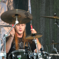 A drummer with long hair and a black shirt is playing a drum set on stage at Kilkim Žaibu 2011. His right hand holds a drumstick hitting a cymbal. The drum set includes a bass drum, toms, and cymbals. He is focused on his performance with studio lighting and music equipment visible in the background.