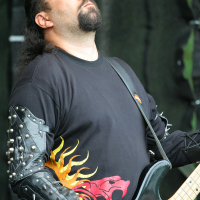 A person with long dark hair, a beard, and mustache is playing a black electric guitar. They are wearing a black shirt with a red and yellow sun design and black armbands adorned with metal spikes. Their eyes are closed as they appear deeply absorbed in the music, reminiscent of Kilkim Žaibu 2011.