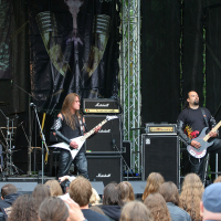 Three musicians perform on an outdoor stage at the 2011 Kilkim Žaibu festival. The guitarist on the left plays a Flying V guitar while the bassist on the right plays a bass guitar. A drummer is visible behind them. The audience, with long hair, stands close to the stage amidst amplifiers and a backdrop with the band logo.